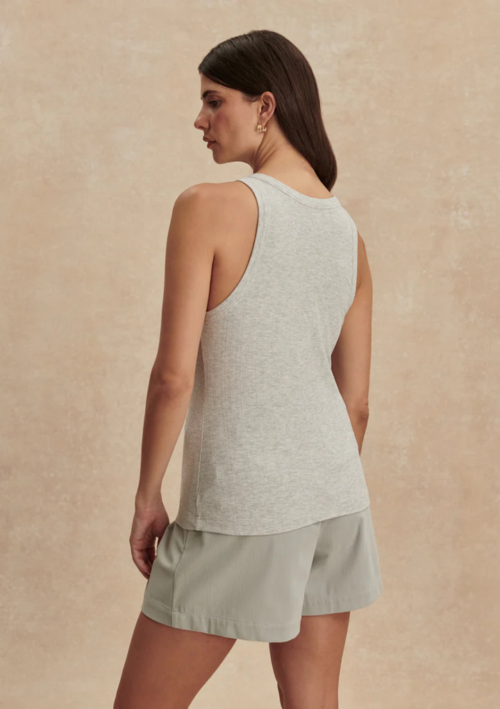 A woman shown from the back wearing a light heather gray sleeveless tank top and light sage green shorts against a textured beige studio backdrop.