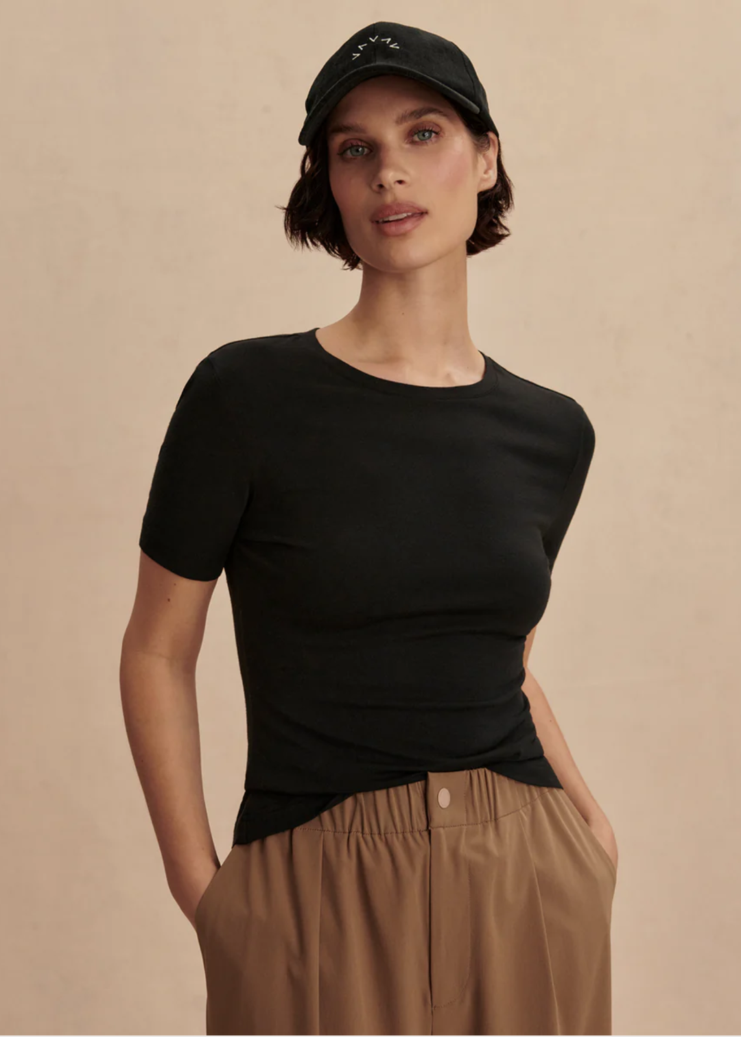 A woman wearing a fitted black short-sleeve crewneck top, tan pleated trousers, and a black baseball cap, standing against a plain beige studio background.