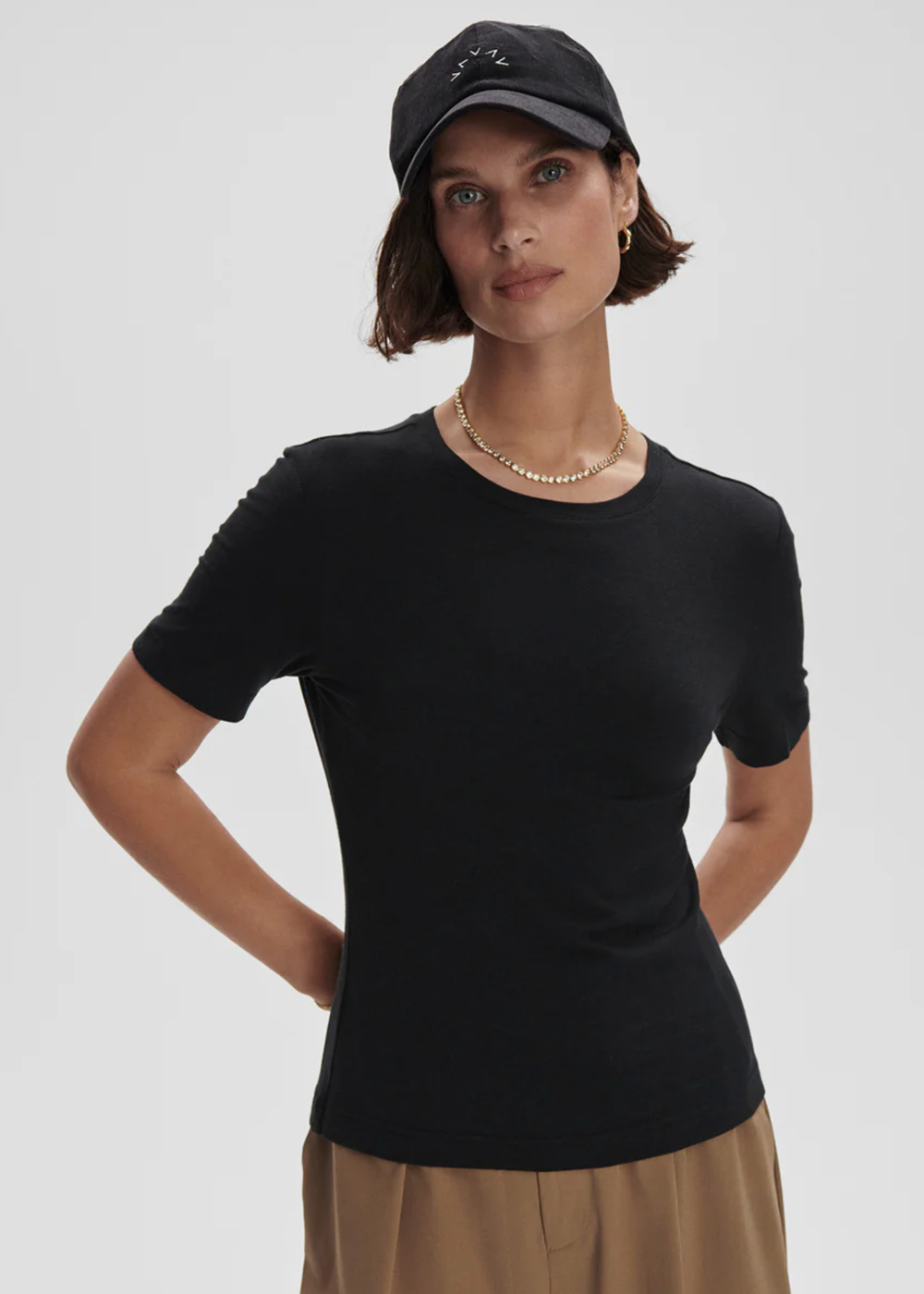 A woman modeling a fitted black short-sleeve crewneck t-shirt, paired with a black baseball cap, gold chain necklace, and beige trousers, against a light gray studio background.