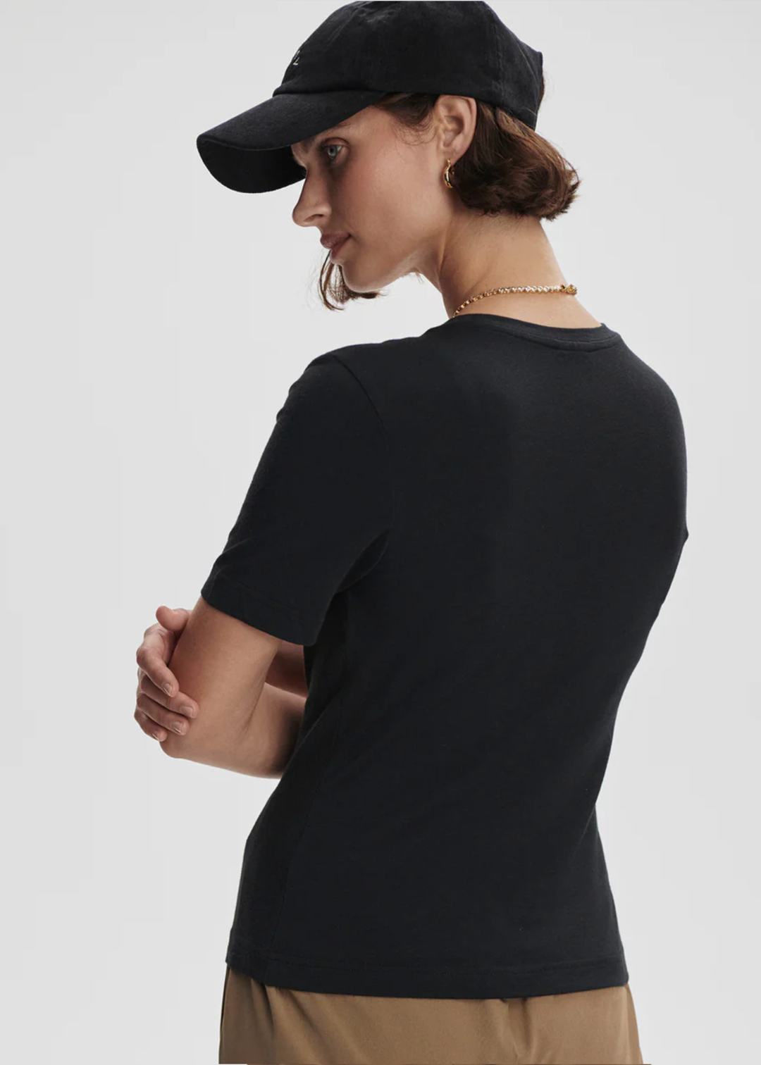 A woman shown from the back wearing a fitted black short-sleeve crewneck t-shirt, a black baseball cap, and tan pants, against a plain light gray studio background.