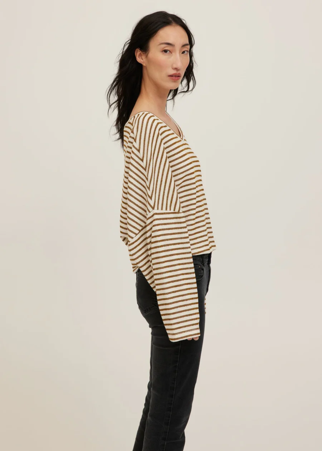 A woman modeling a beige and brown horizontally striped long-sleeve top with black jeans, shown from a side profile against a plain light background.