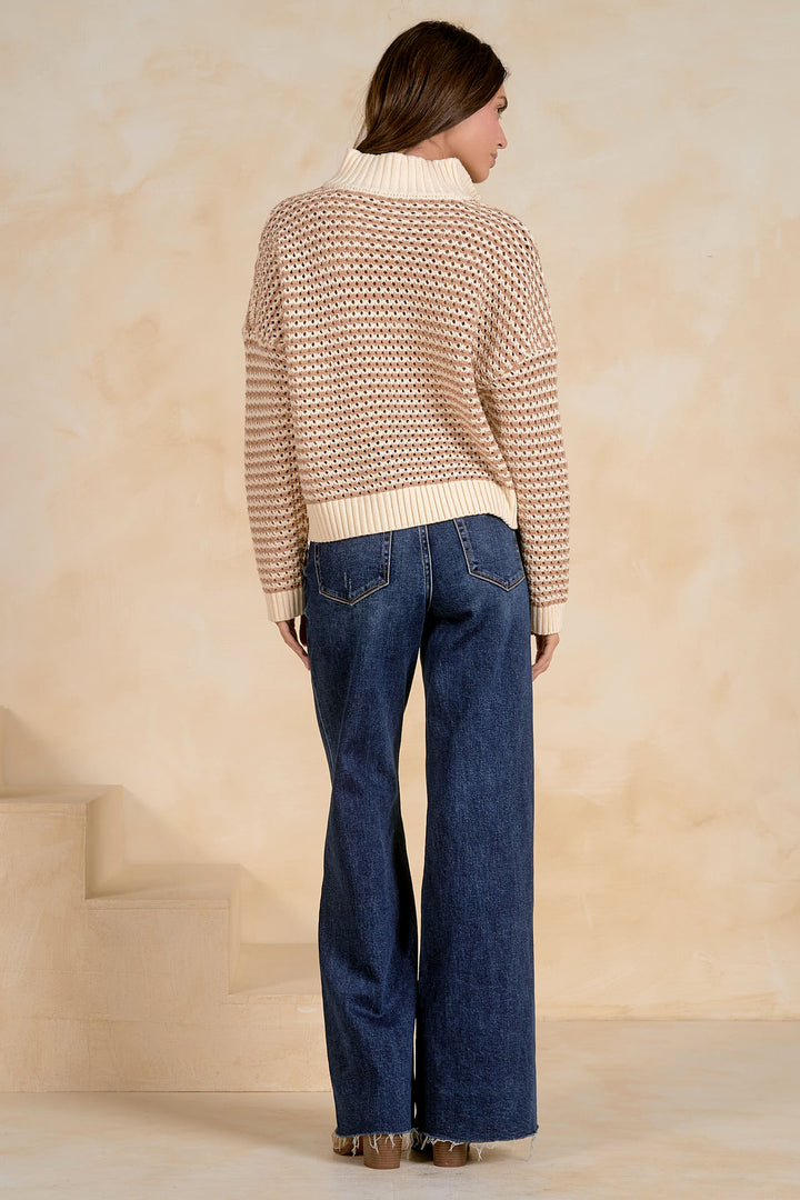 Woman wearing a patterned sweater and blue jeans against a beige wall.