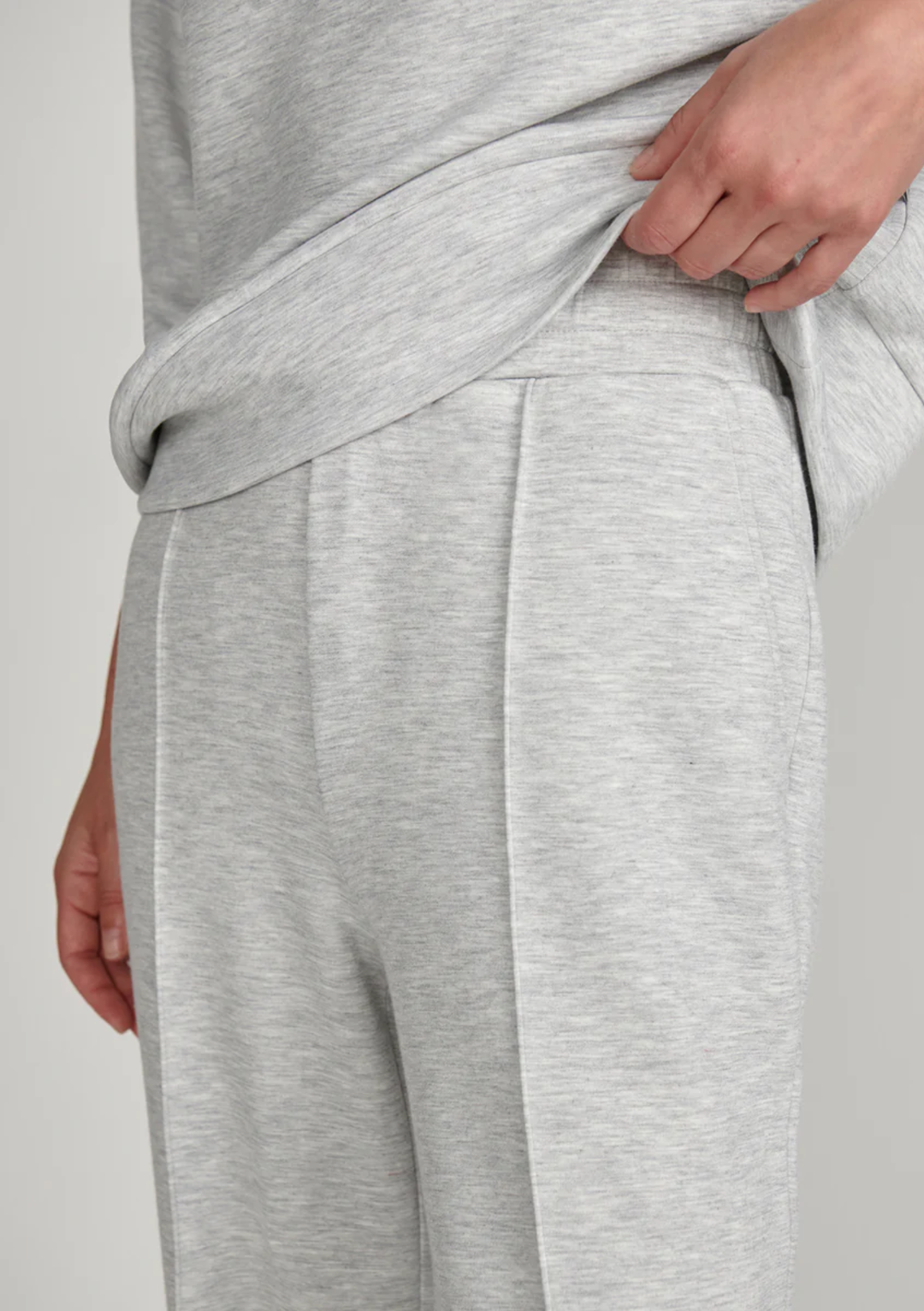 Close-up of a person wearing light heather gray lounge pants, pulling the waistband slightly upward with one hand.