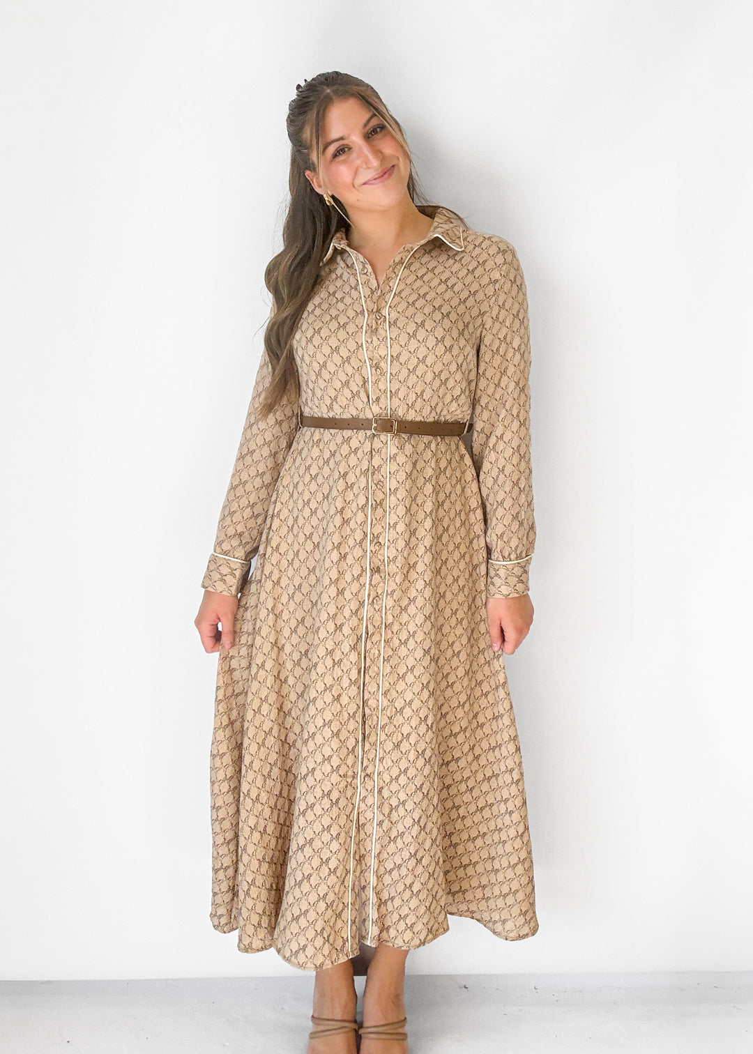 A woman modeling a long-sleeve collared midi dress with a brown geometric pattern on a beige background, featuring a matching brown belt, standing against a plain white wall.