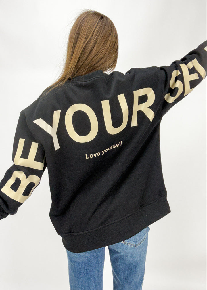 Person wearing a black hoodie with 'Be Your Self' text in gold on a white background