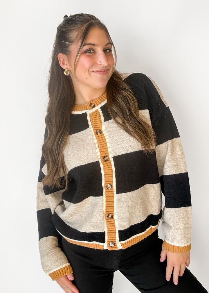 A woman wearing a knit cardigan sweater with wide horizontal stripes in black, cream, and mustard yellow, featuring a mustard yellow button placket and ribbed trim.