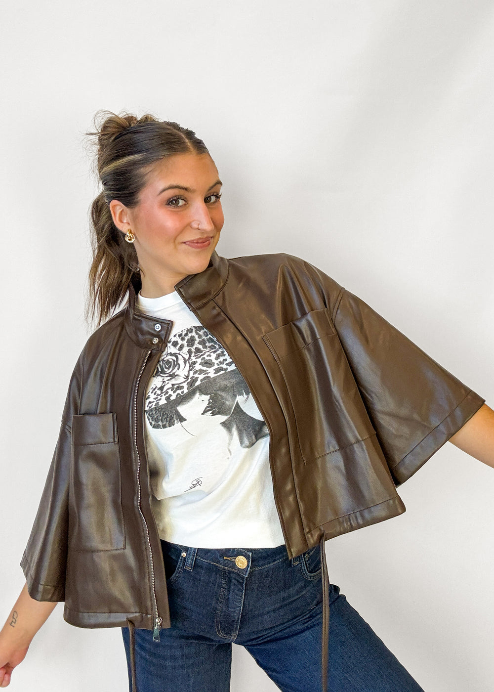 A woman wearing a brown faux-leather cropped jacket with wide short sleeves, layered over a white graphic t-shirt and blue jeans, posing against a plain white studio background.