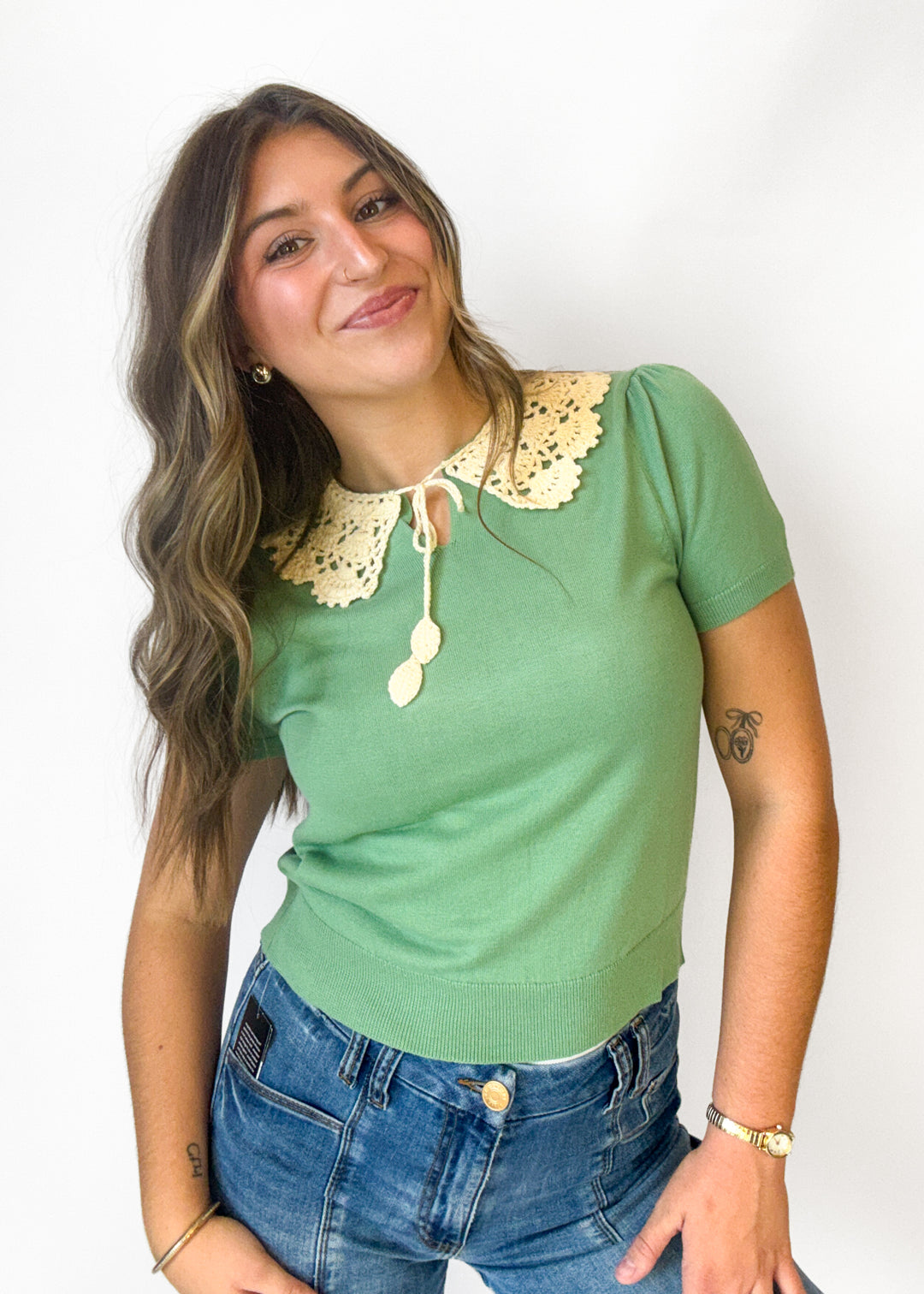 A woman wearing a sage green short-sleeve knit top with a cream-colored crochet n collar and a tie detail, paired with blue jeans, against a plain white background.