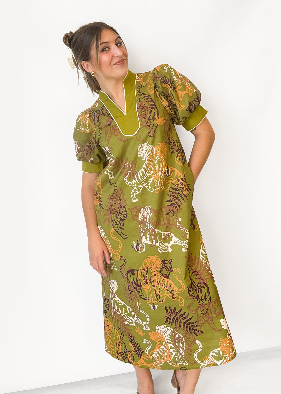 A woman modeling a short-sleeved midi dress with a green base and a bold tiger and leaf print in orange, brown, and white, set against a plain white studio background.