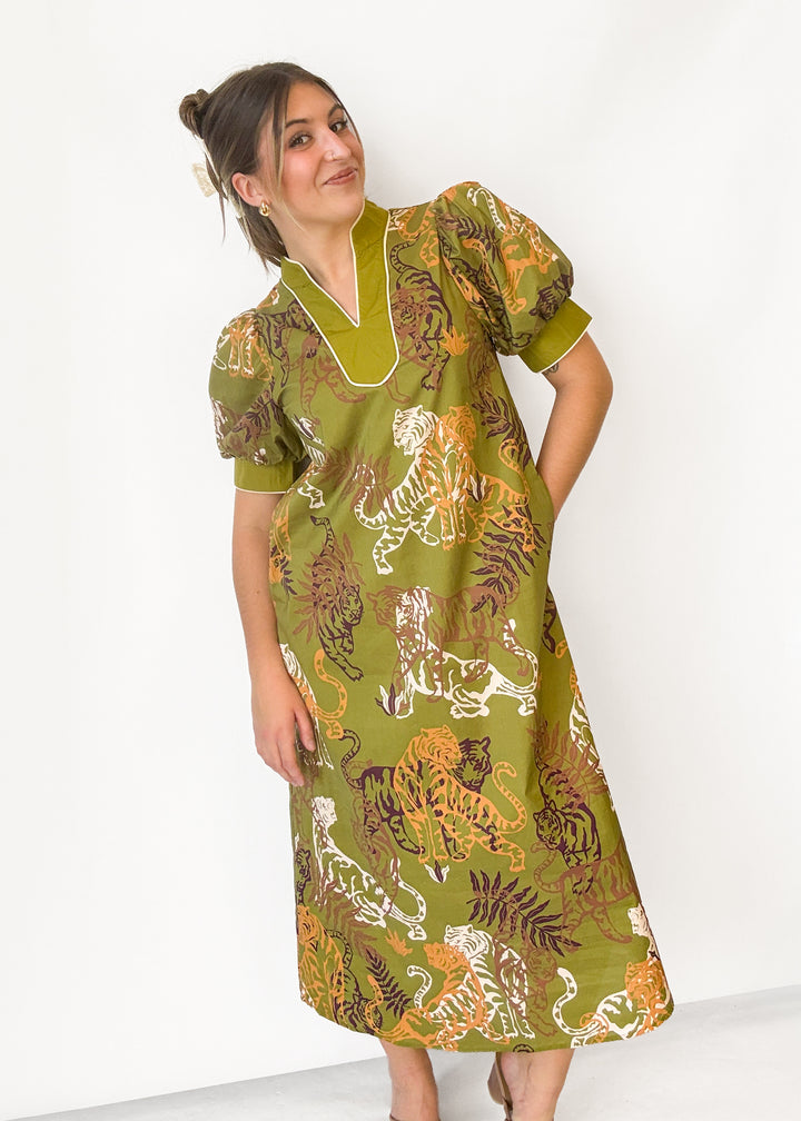 A woman modeling a short-sleeved midi dress with a green base and a bold tiger and leaf print in orange, brown, and white, set against a plain white studio background.