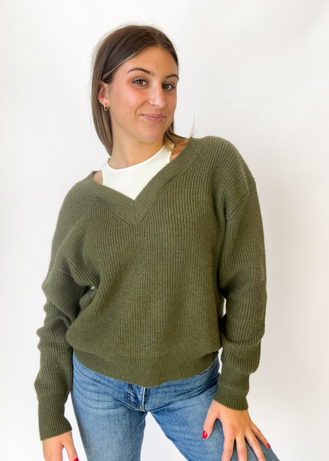 A woman wearing an olive green ribbed knit sweater over a white t-shirt and blue jeans, standing against a plain white background.