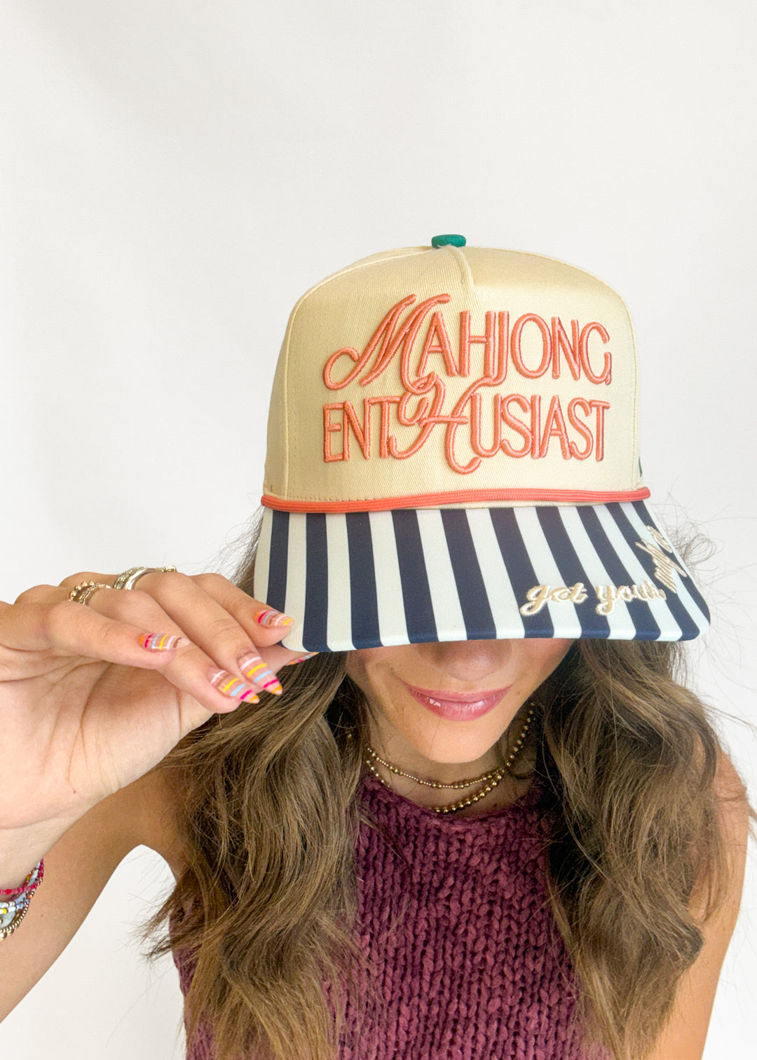 A woman wearing a beige snapback cap with orange embroidered text reading 'MAHJONG ENTHUSIAST' and a navy-and-white striped brim, holding the brim over her eyes. The photo is cropped from the chest up against a plain light background.