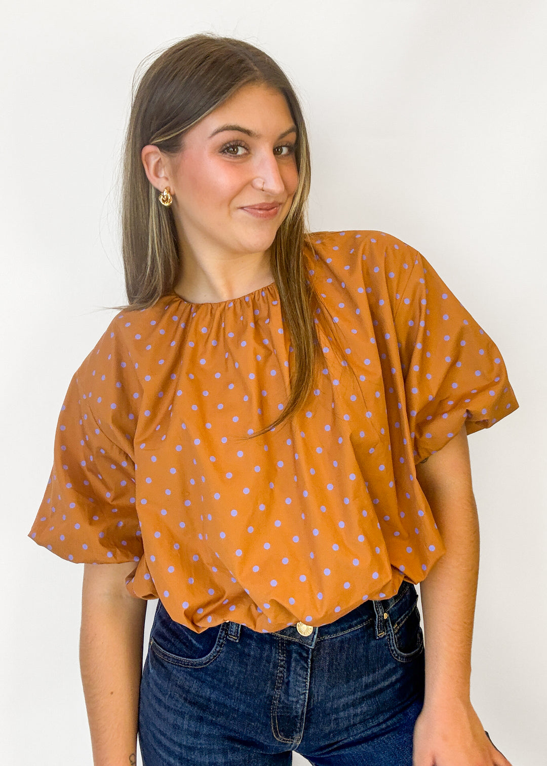 A woman wearing a mustard orange short-sleeve blouse with light blue polka dots and dark blue jeans, standing against a plain white background.