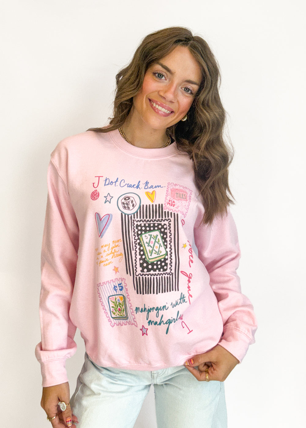 A woman wearing a light pink crewneck sweatshirt with colorful graphic prints and text, paired with light blue jeans, posing against a plain white background.