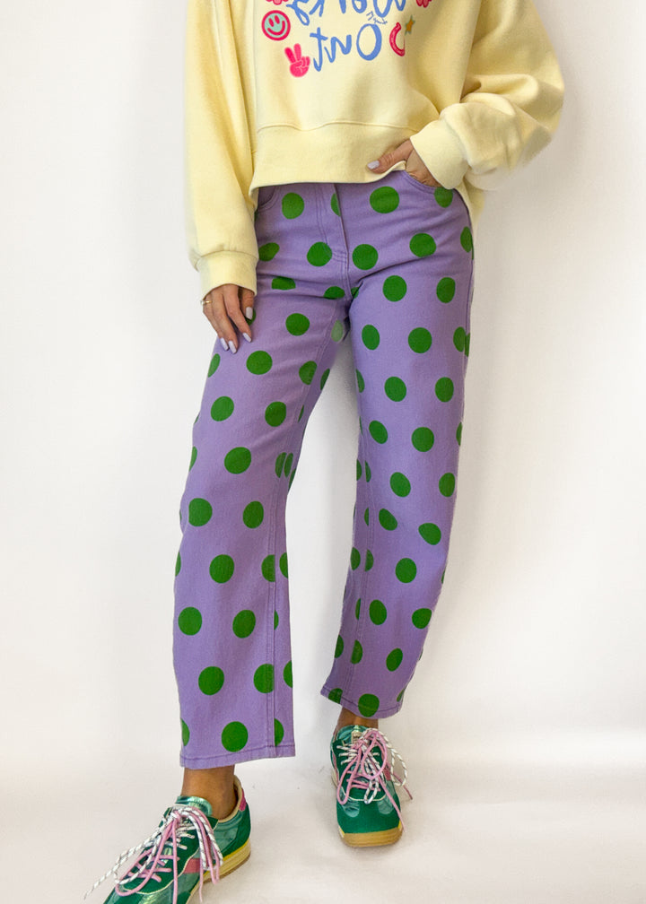 Person wearing a yellow sweatshirt with colorful text, purple pants with green polka dots, and green sneakers on a white background.