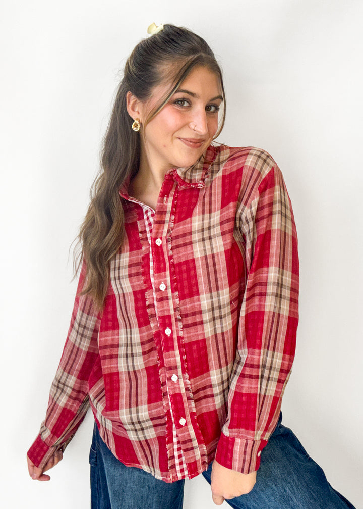 A woman wearing a red and beige plaid long-sleeve button-up shirt with blue jeans, posing against a plain white wall.