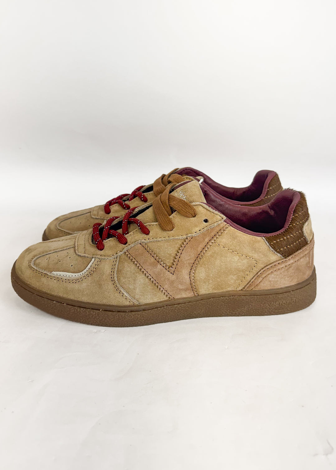 A pair of low-top sneakers in beige suede and tan leather, featuring brown laces with red tips, a brown sole, and a burgundy interior, shown against a plain white background.