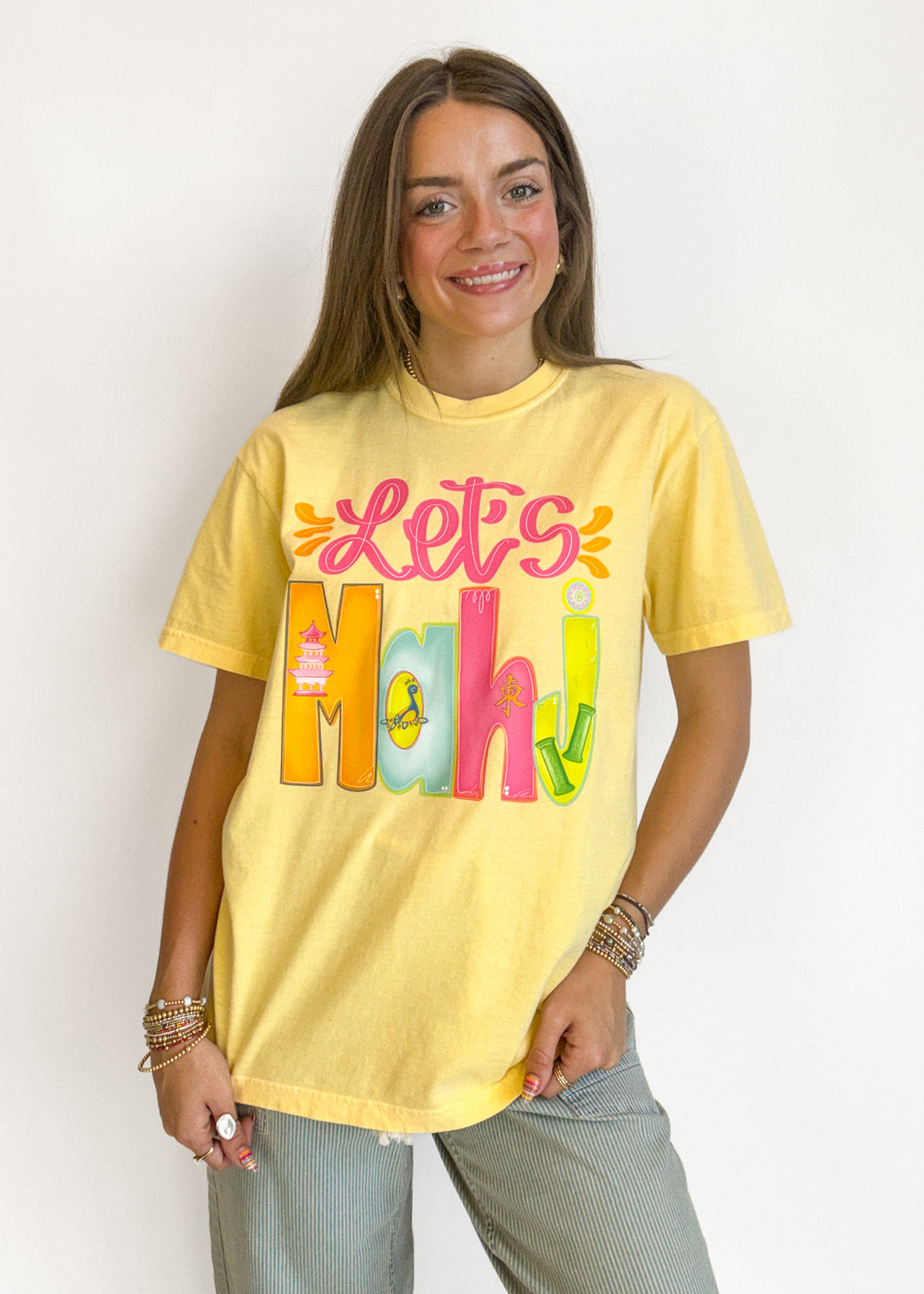 A woman wearing a yellow graphic t-shirt with 'Let's Maki' printed in colorful bubble letters, paired with light gray pants, against a plain white background.
