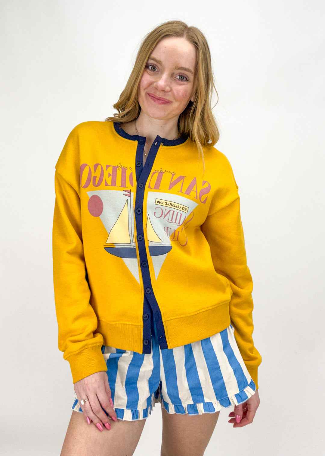 Woman wearing a yellow sweatshirt with a graphic design and blue and white striped shorts on a white background